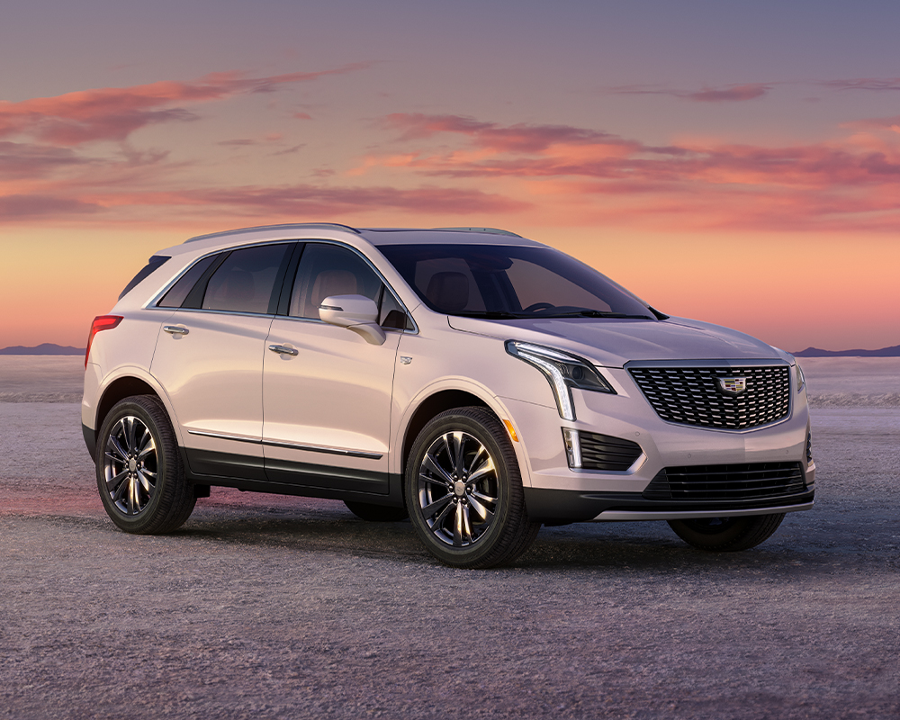A white Cadillac XT5 parked on a flat, open desert landscape under a vibrant pink and orange sunset sky.